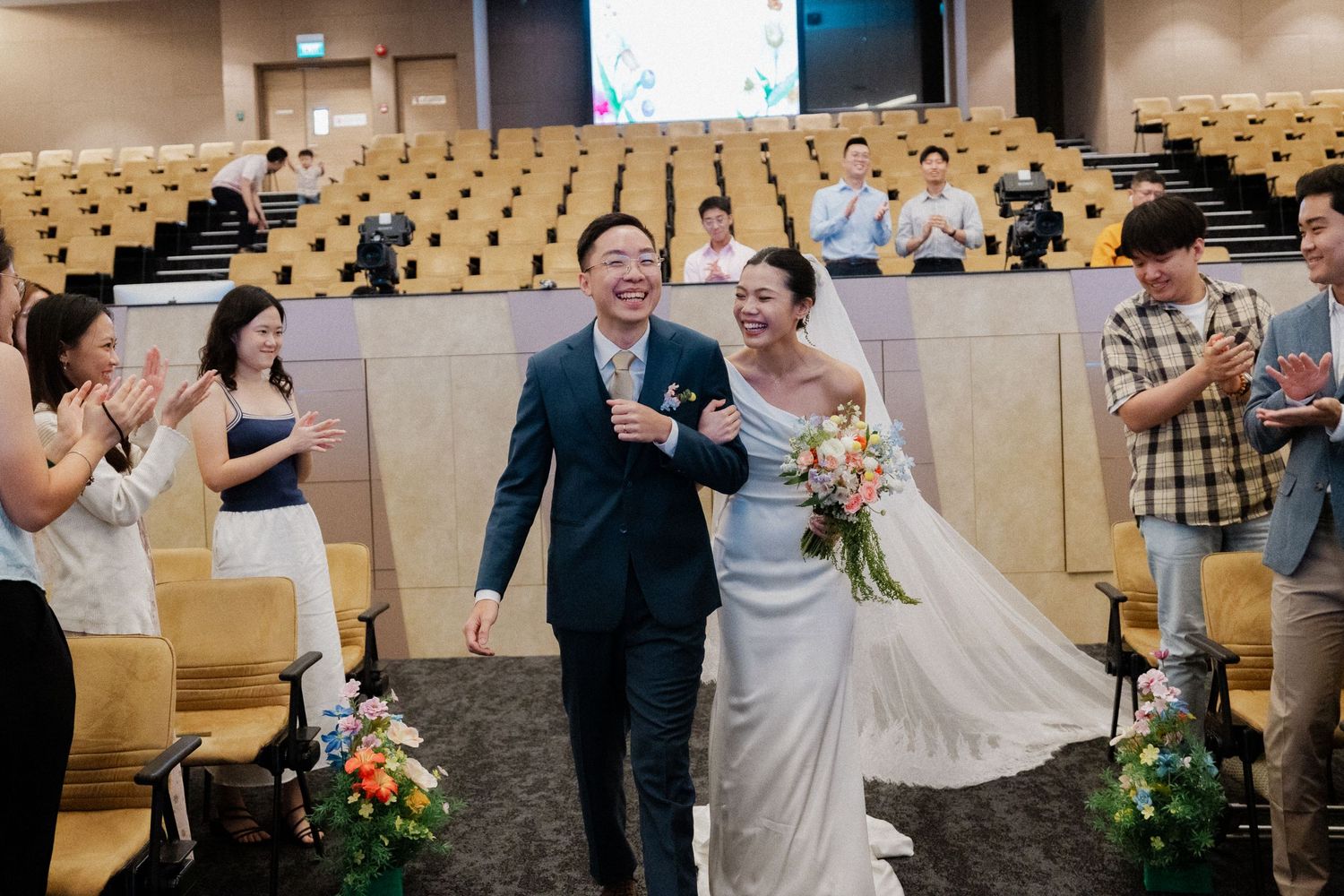 Happy newlywed Asian couple walks down the aisle at their wedding ceremony in a modern venue.