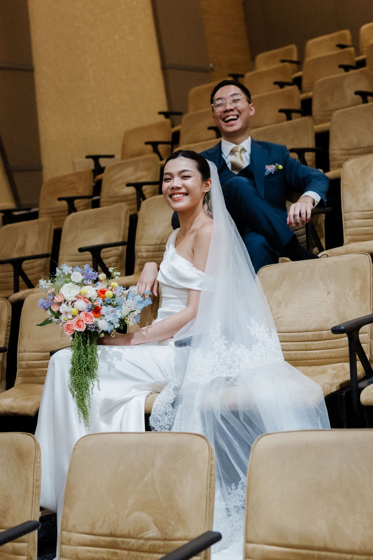 Smiling bride and groom sitting in auditorium seats, bride holding colorful wedding bouquet with veil.