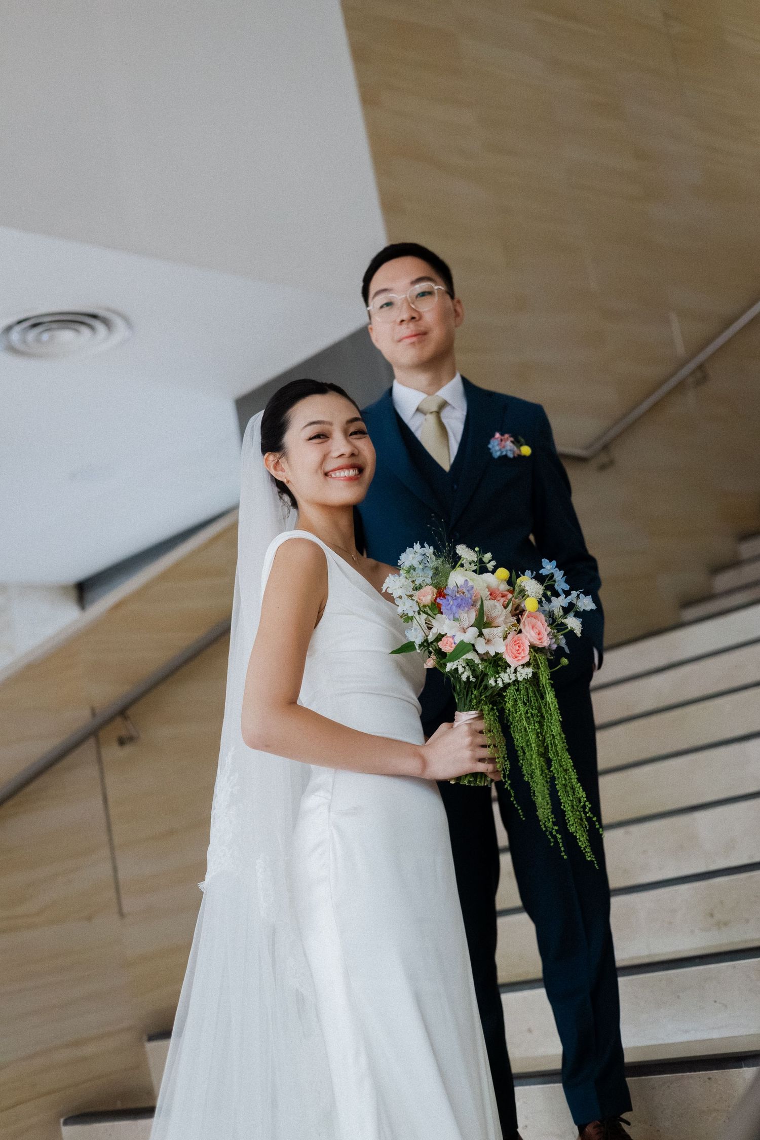 Asian bride and groom pose on marble staircase, bride holding colorful bouquet with trailing greenery, groom in navy suit.