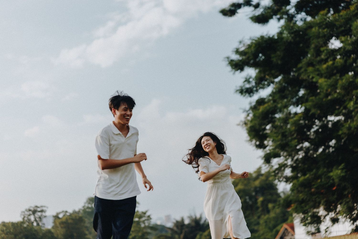 Couple running playfully in a lush green park setting with trees in the background on a sunny day.