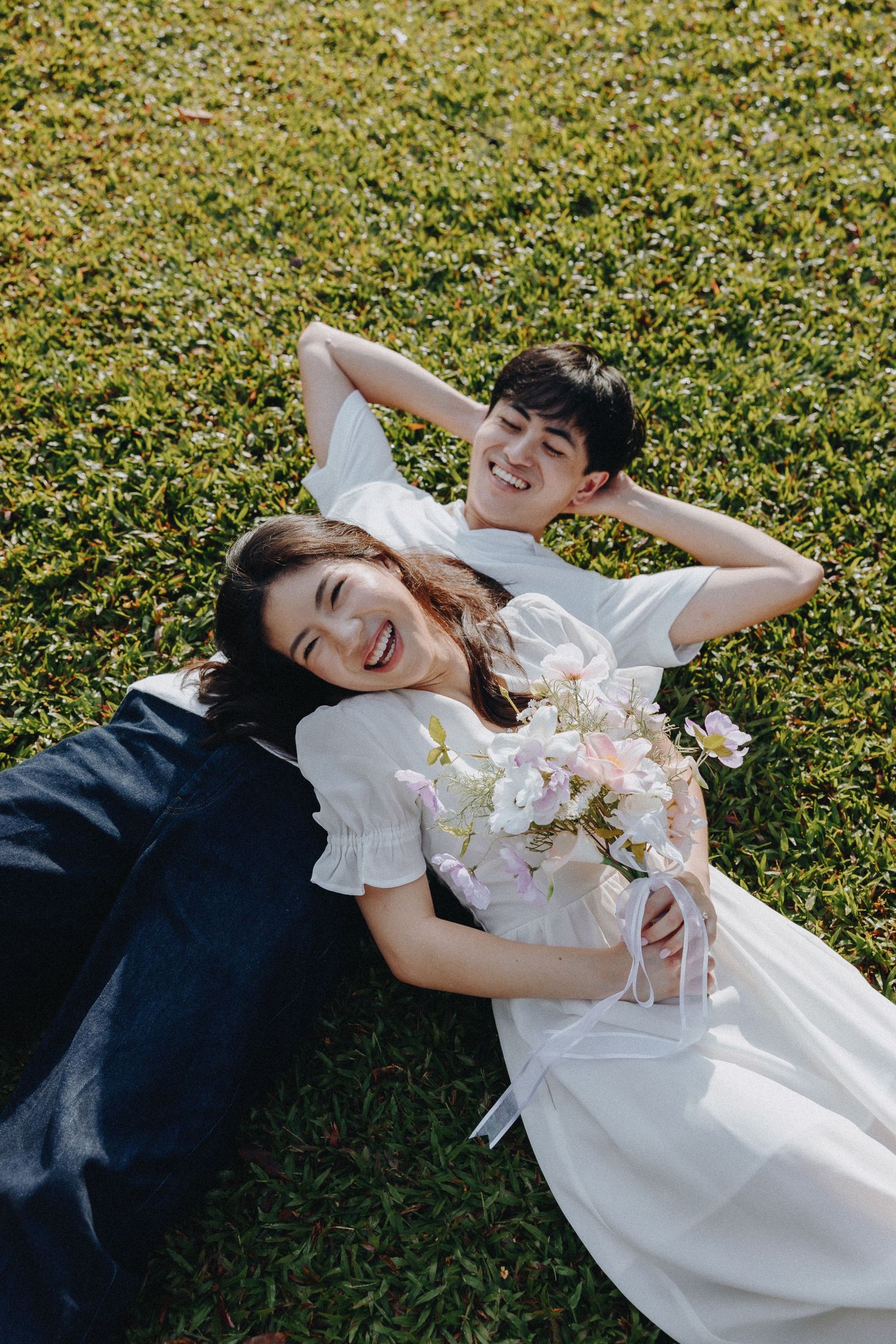 A couple lies on grass with flowers laughing together during a photoshoot.