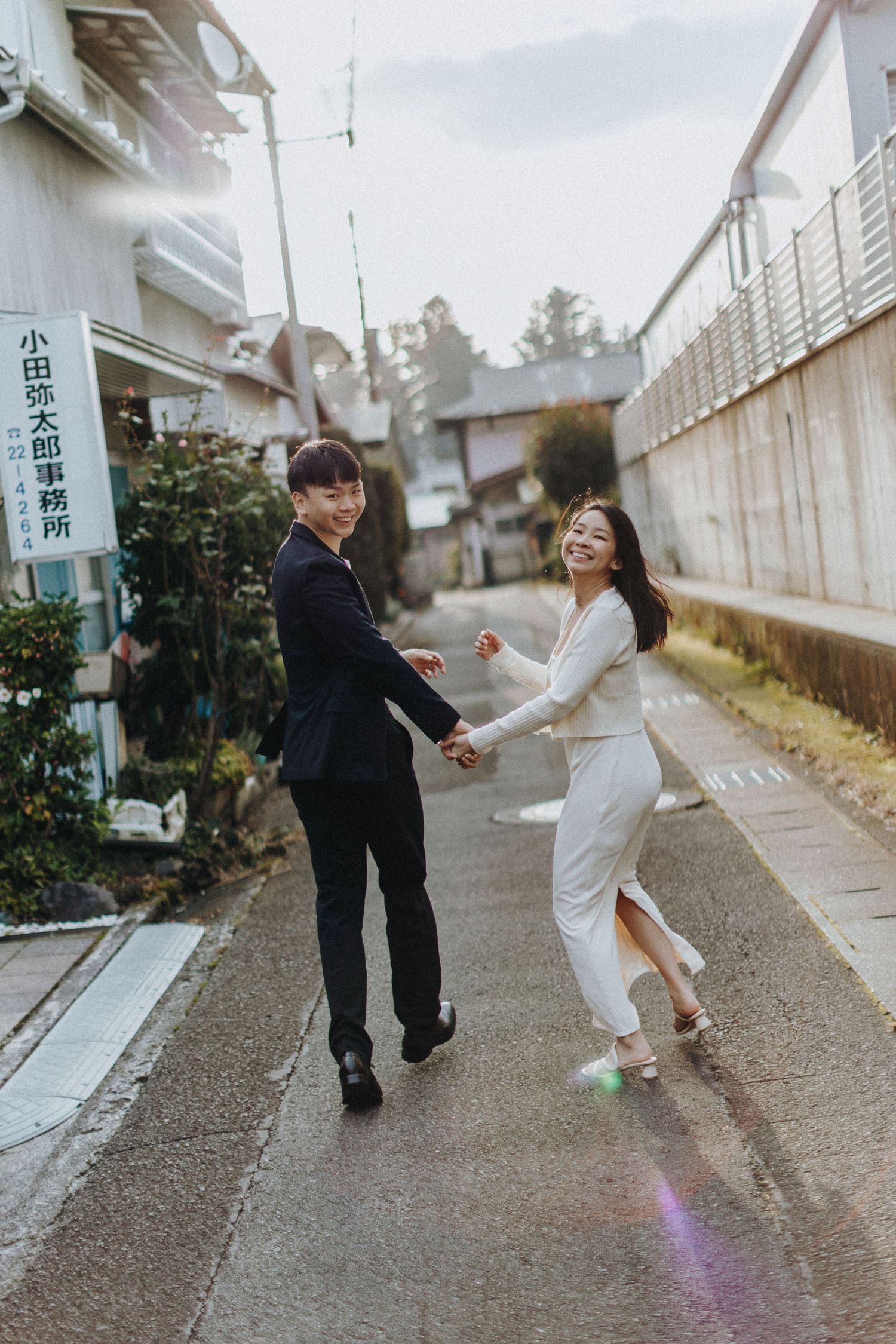A couple walking and dancing along a narrow urban pathway with buildings in the background.