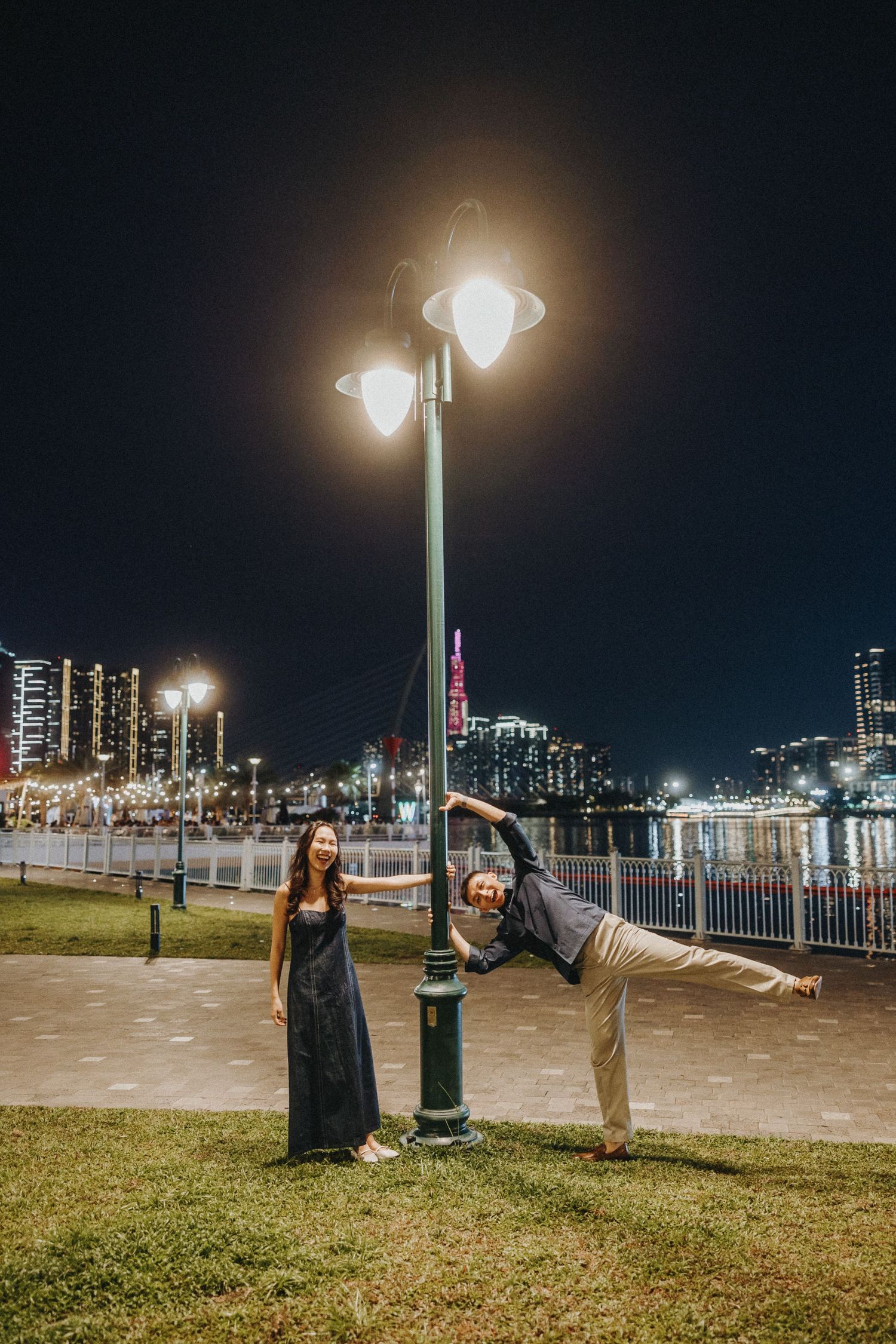 Couple posing playfully by illuminated lamp post at night along waterfront with city skyline in background.