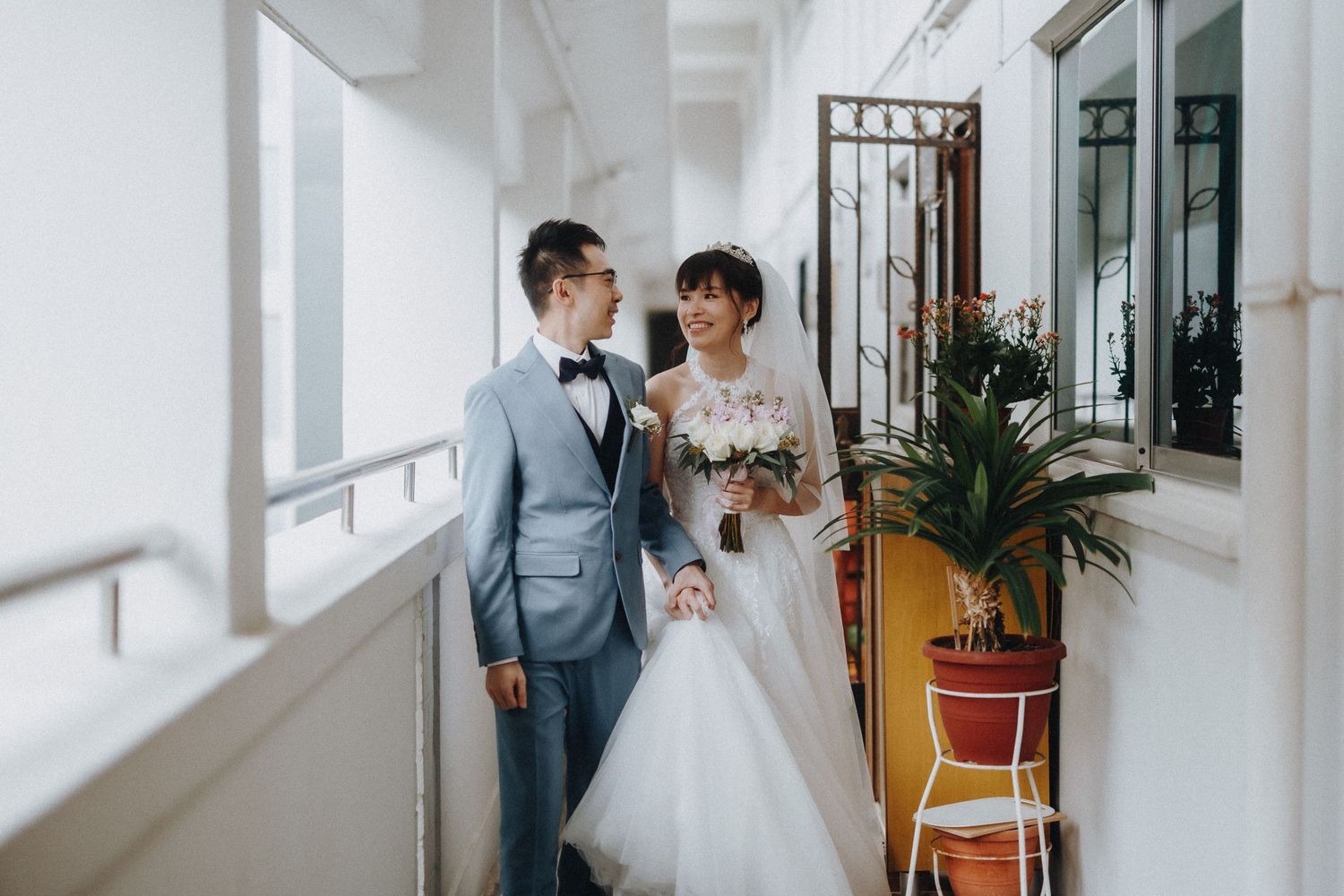 A newlywed couple poses in an elegant white hallway with a potted plant on a yellow chair.