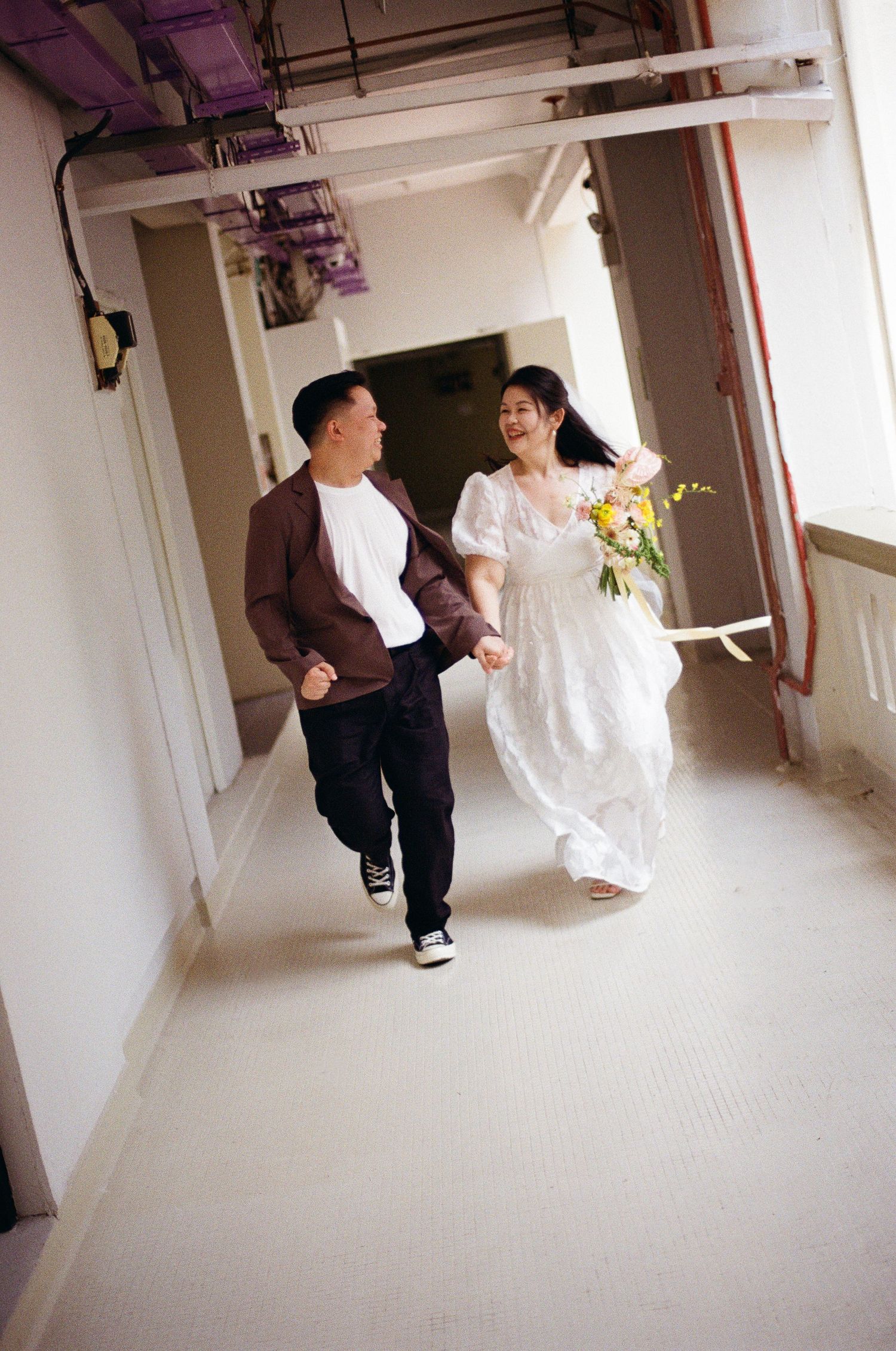 A couple walks together laughing in a white corridor while holding flowers.