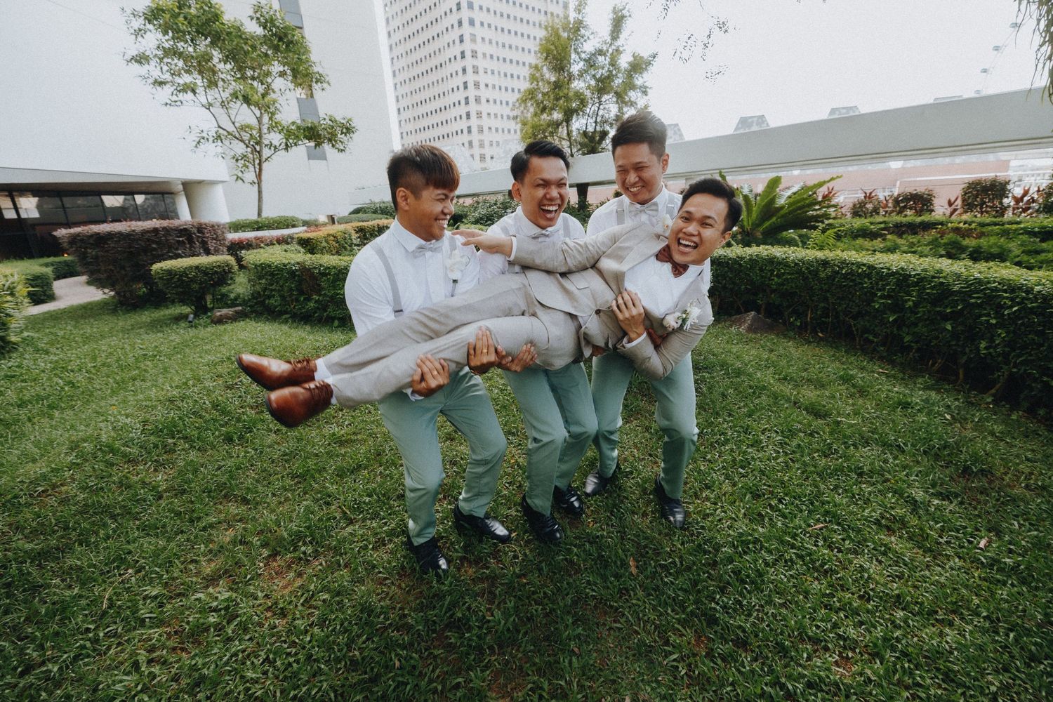 Groomsmen in matching outfits playfully lifting someone in garden setting.