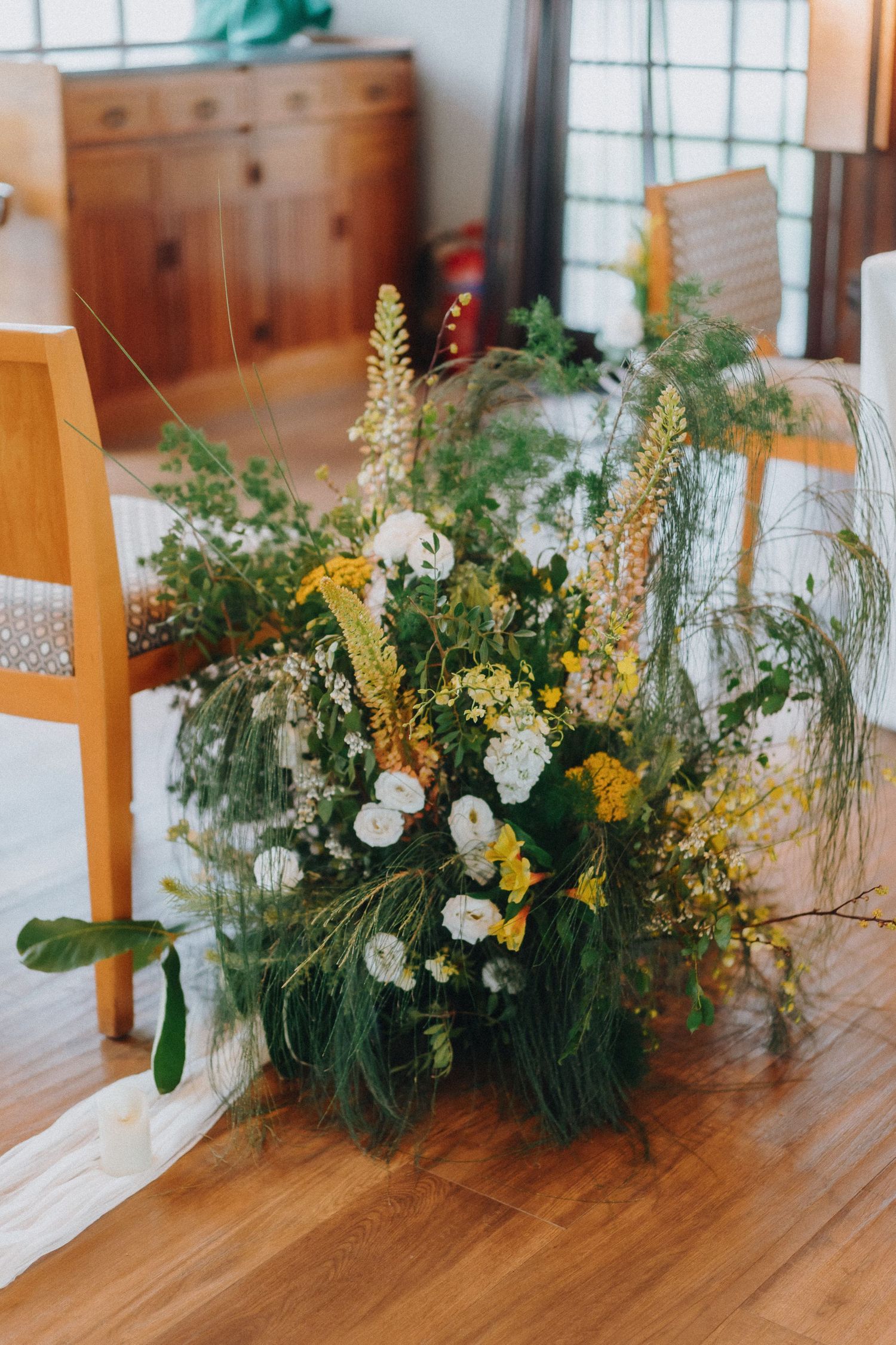 A wildflower arrangement with yellow blooms and greenery sits on a wooden floor near chairs.
