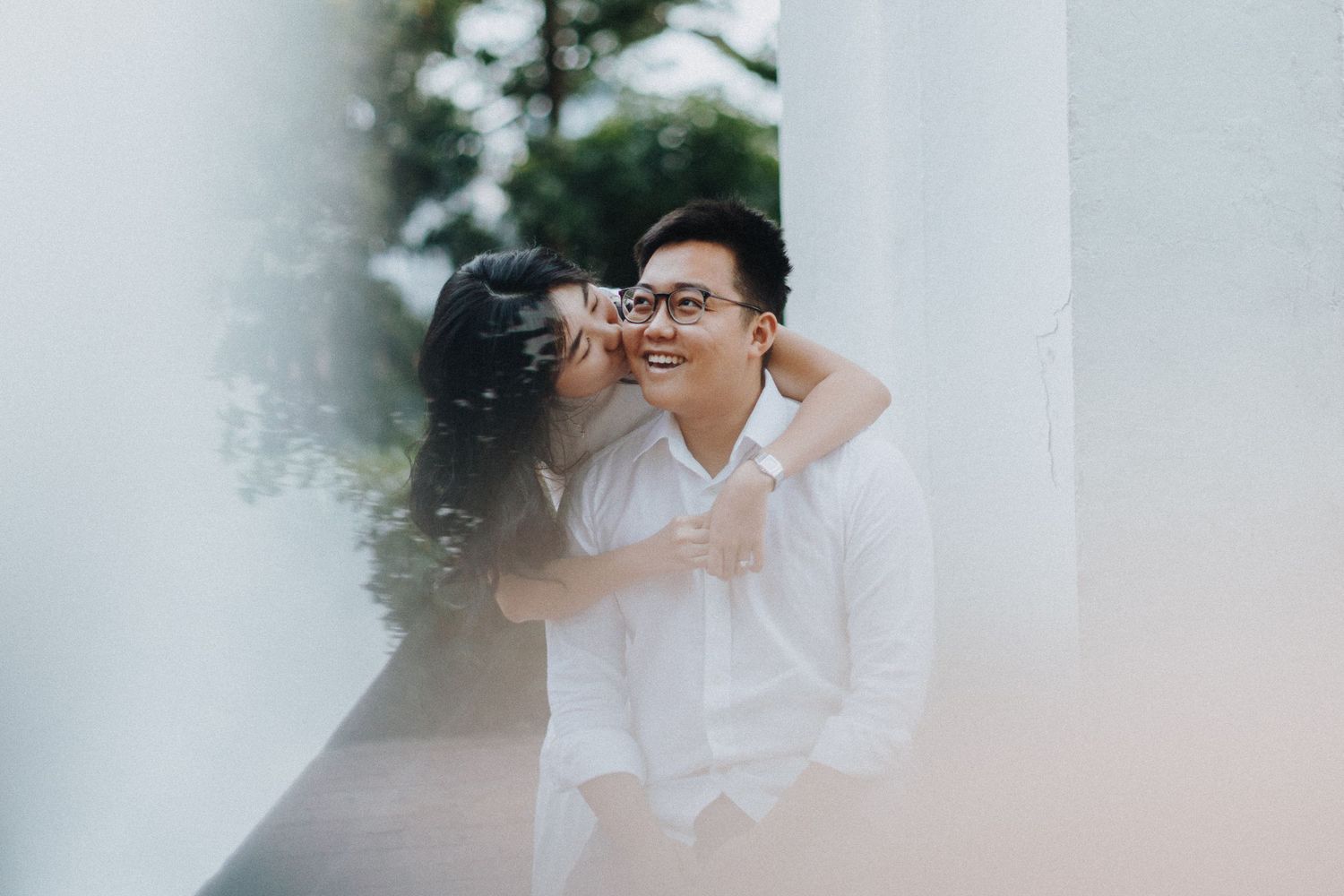 A romantic couple in white share an intimate moment against a dreamy white backdrop with greenery.