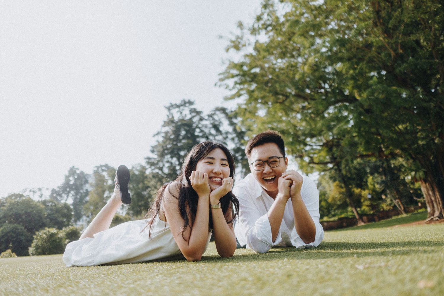 A couple lies on grass outdoors making playful expressions during a photoshoot.