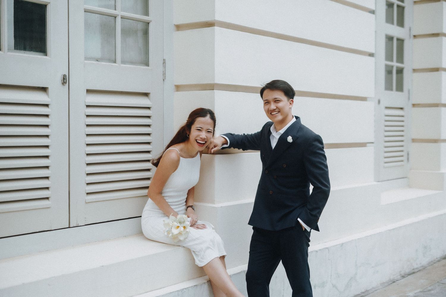 A couple poses playfully against a white architectural backdrop in formal attire.