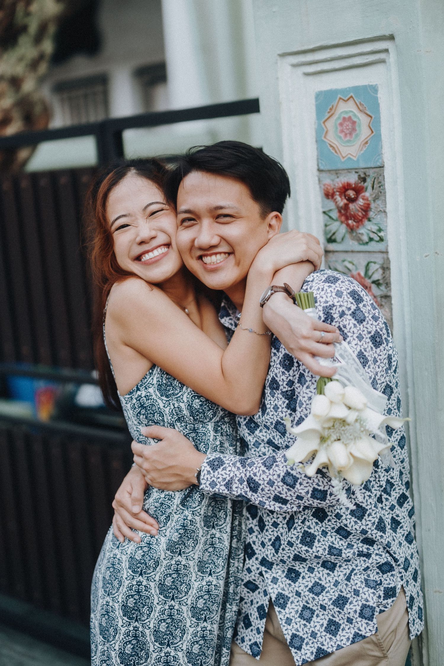 A couple wearing matching blue and white patterned outfits embrace and smile while standing against a wall.