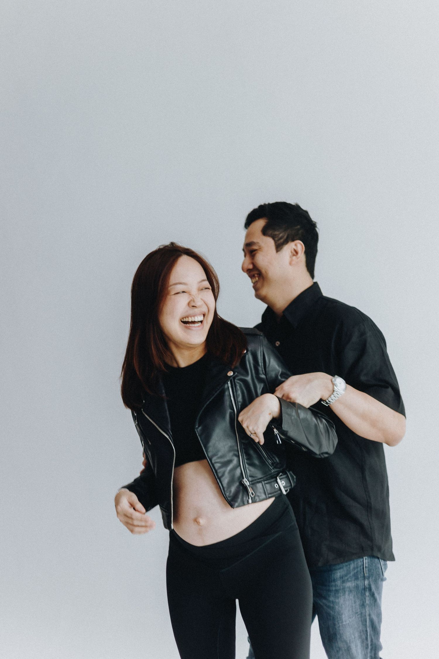 Joyful couple shares a candid moment during pregnancy photoshoot wearing black outfits against white background.