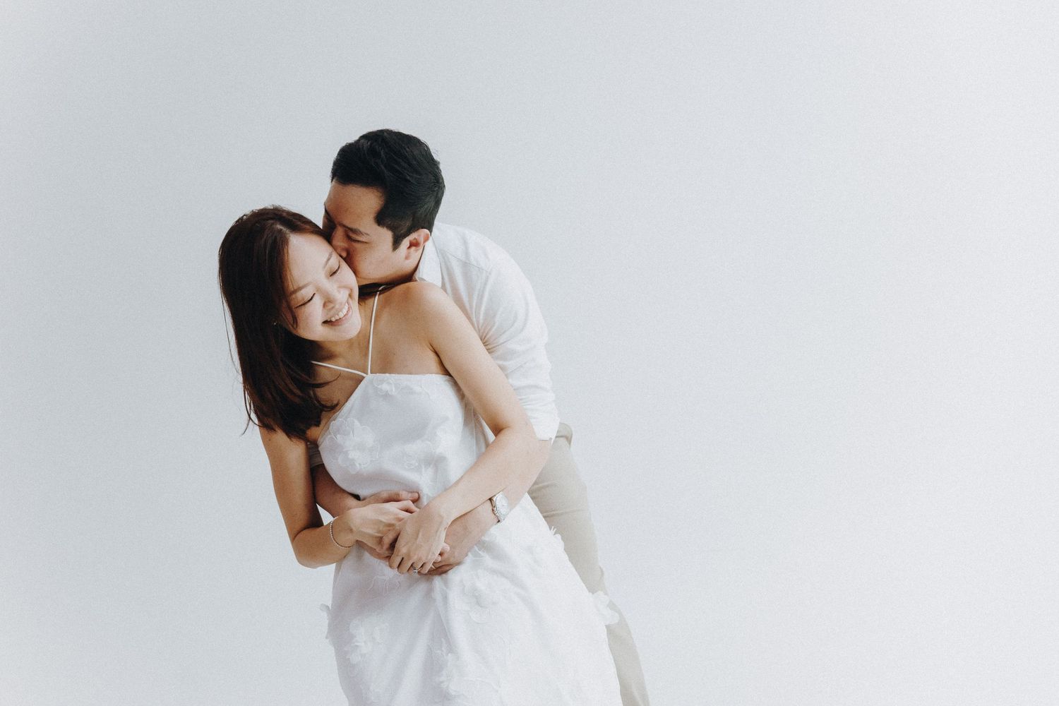 Two people in white attire embrace intimately against a minimalist white background.