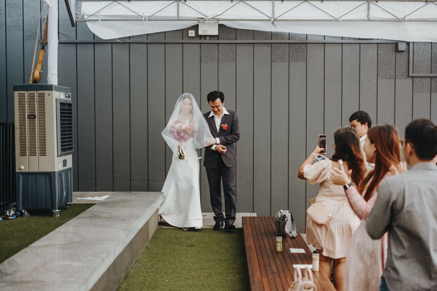 Wedding couple poses for ceremony photos while guests capture moments on their phones on an outdoor rooftop venue.