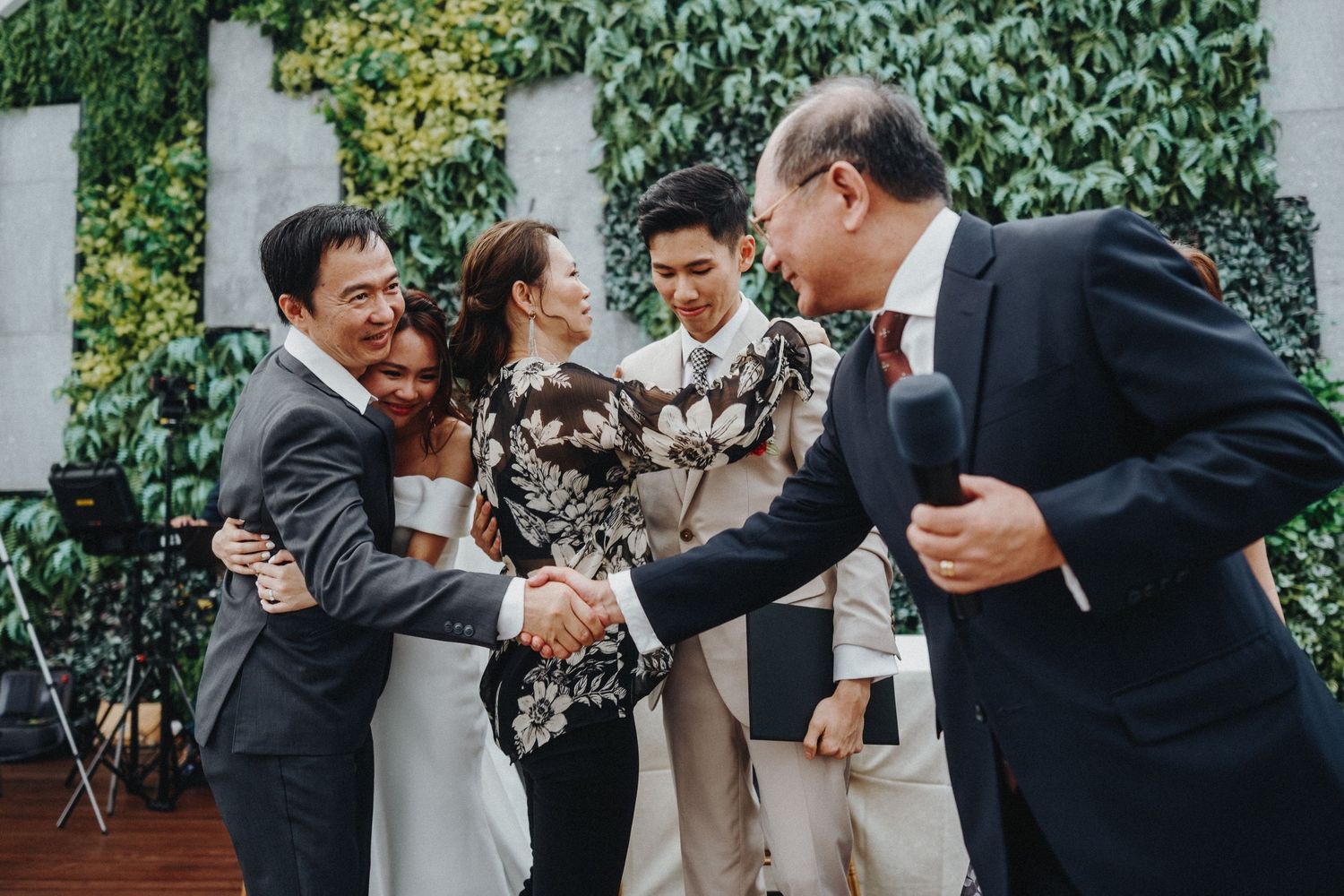 A group in formal attire sharing laughter and handshakes at an elegant outdoor venue with ivy-covered walls.