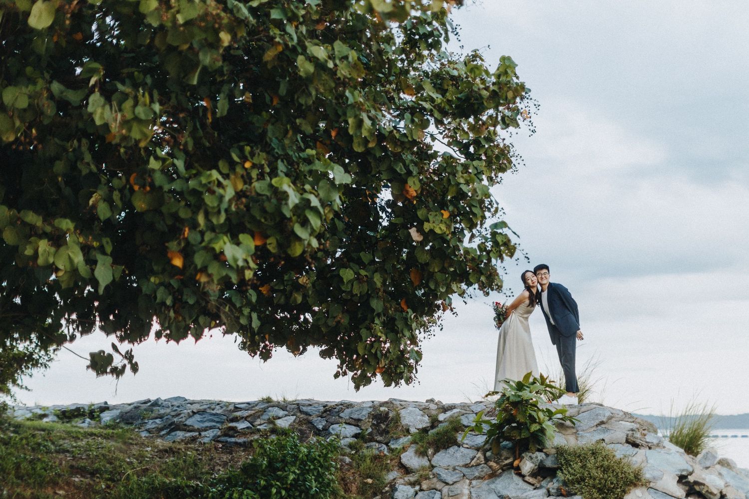 Two people stand on rocky cliff overlooking water, framed by green tree branches against cloudy sky.