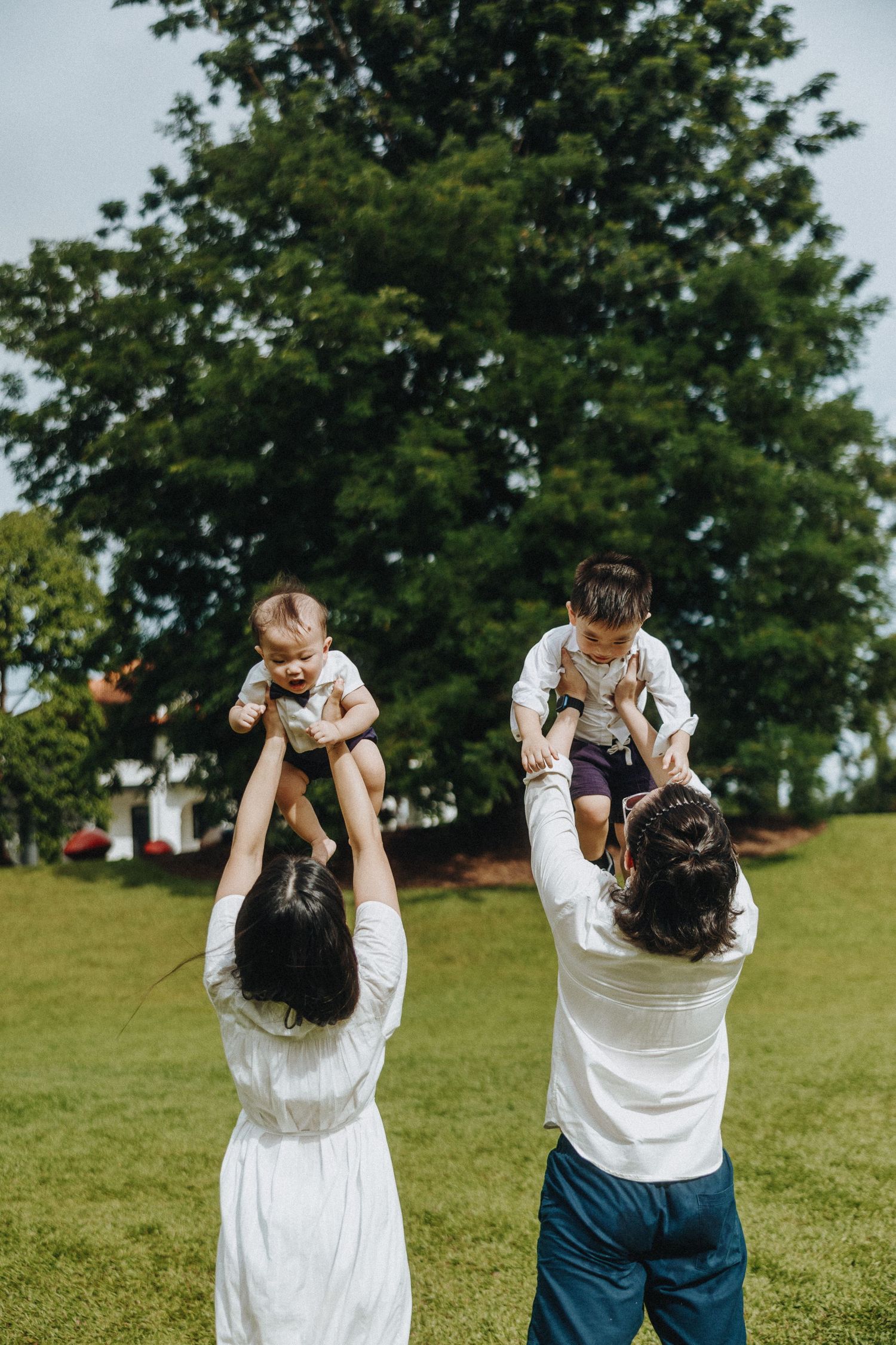 Two people lifting children on their shoulders while playing on a grassy lawn with trees behind them.
