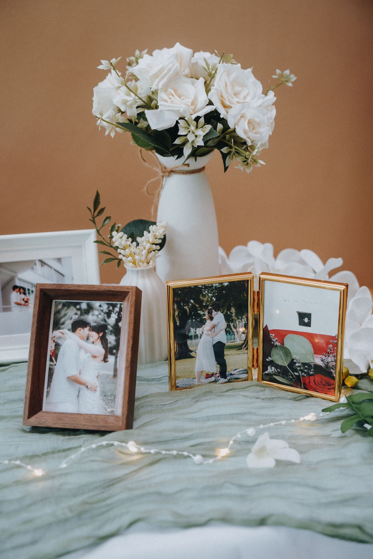White roses in a vase alongside framed photos on a tablecloth.