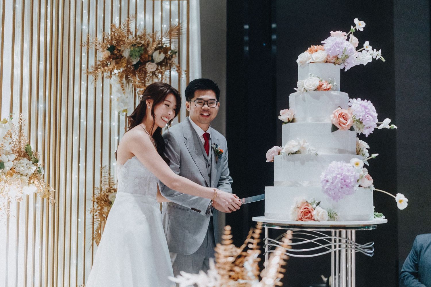 A newlywed couple cuts into an elegant four-tier white wedding cake decorated with pink and purple flowers.