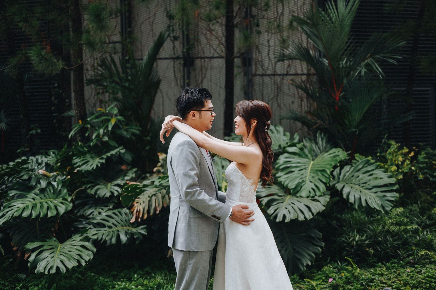 A bride and groom embrace among lush tropical plants and Monstera leaves in an intimate garden setting.