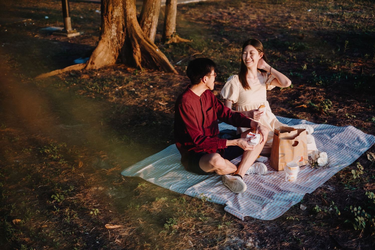 Romantic outdoor picnic scene with couple sitting on white blanket under trees.