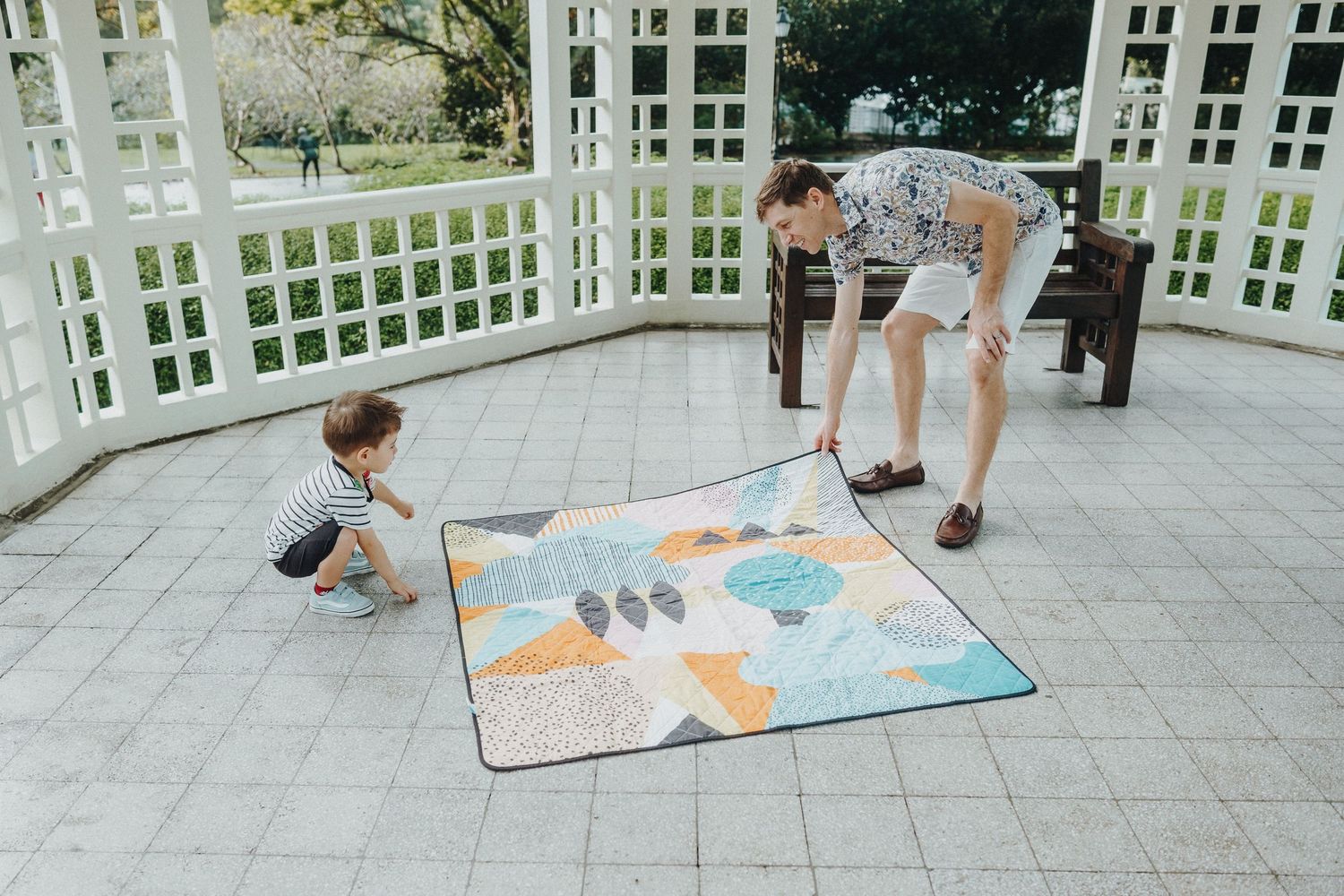 Two people play together on a colorful activity mat in an outdoor gazebo on a sunny day.