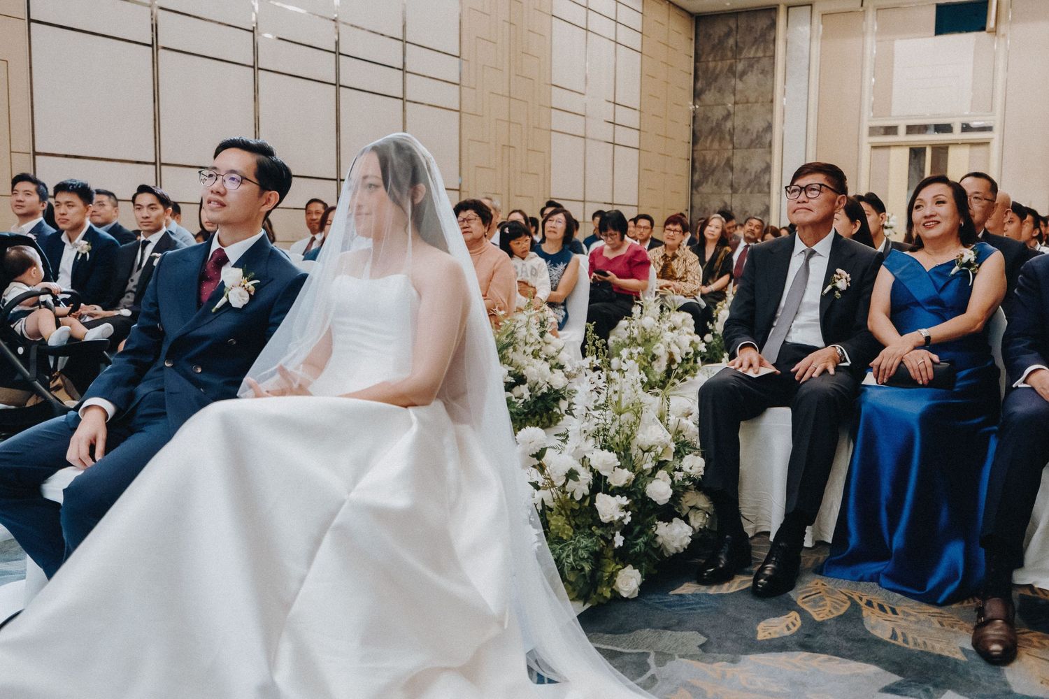 Wedding ceremony in a grand hall with white floral decorations and seated guests.