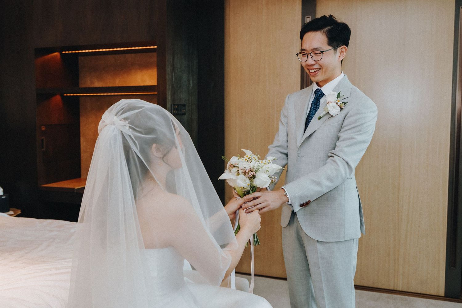 A romantic wedding photoshoot showing a couple in a light-filled room with the bride in white and groom in gray suit.