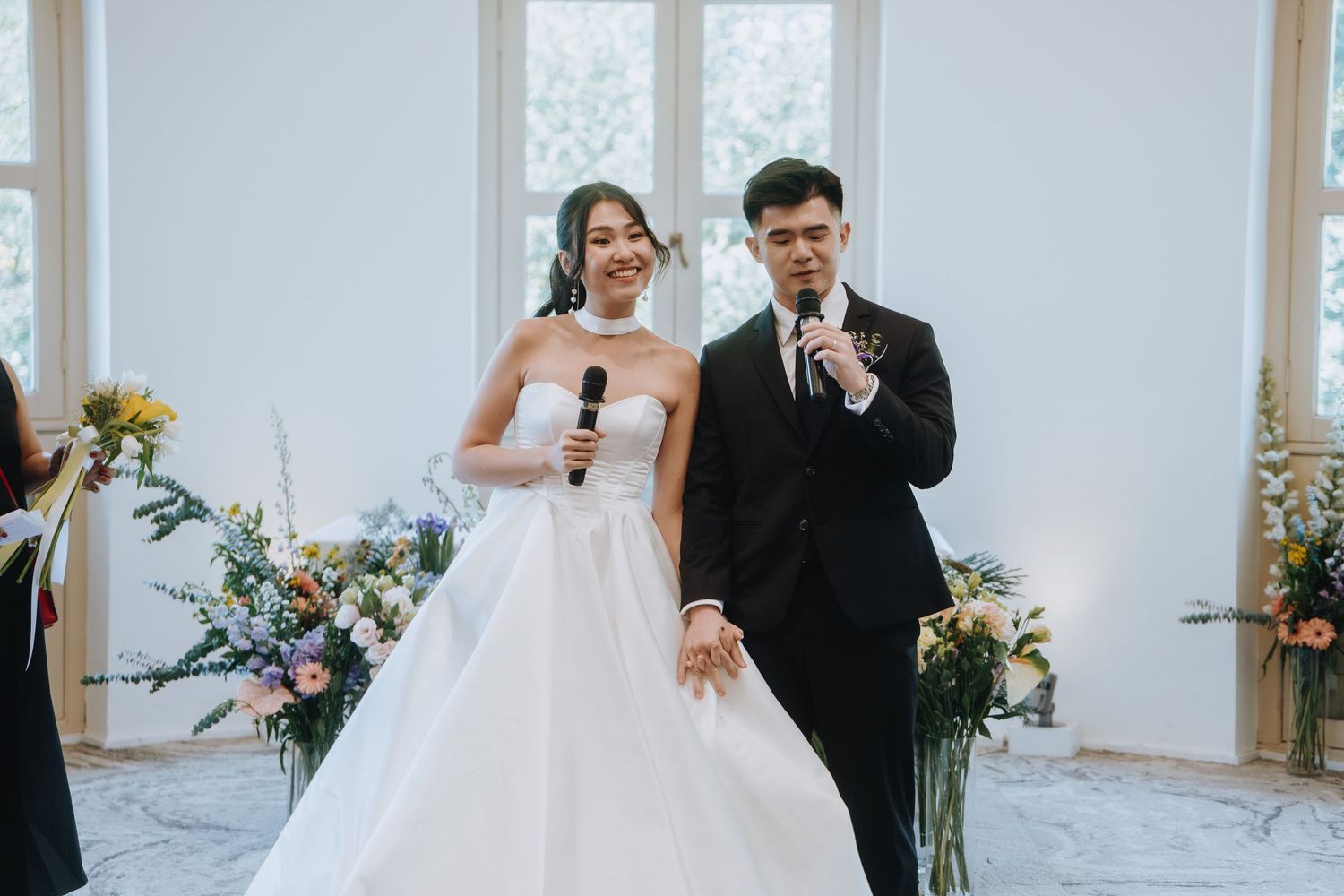 Bride and groom exchanging vows at elegant wedding ceremony, holding hands with floral arrangements behind them.