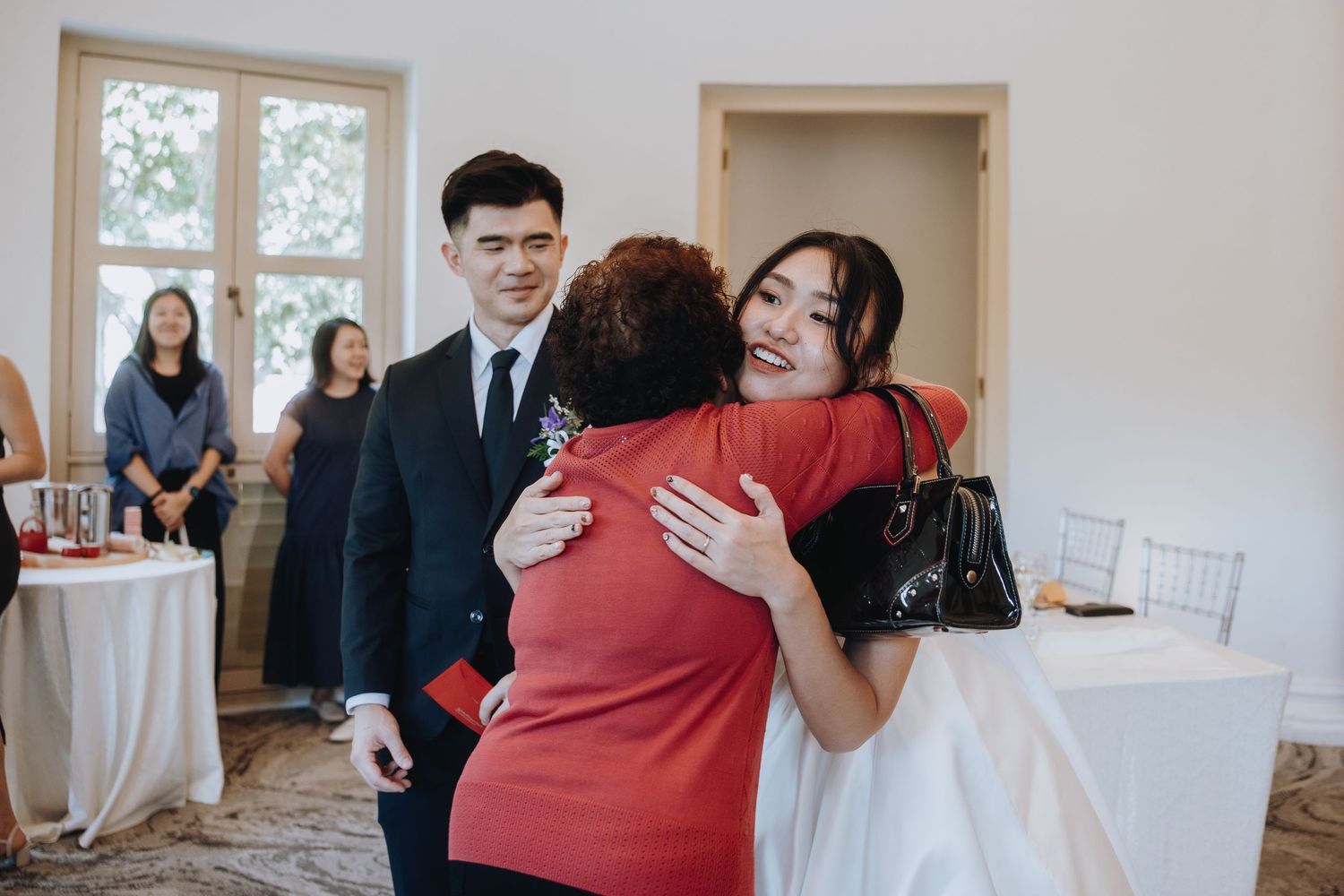 Bride in white dress hugs a guest in red while groom stands smiling at an elegant wedding reception.