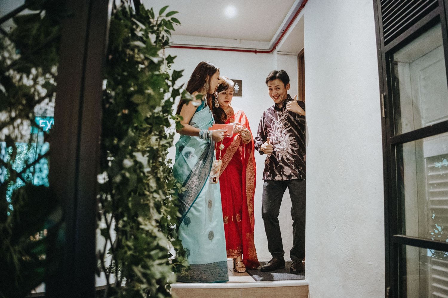 Three people in traditional attire stand on steps, examining fabric together in an elegant indoor setting.