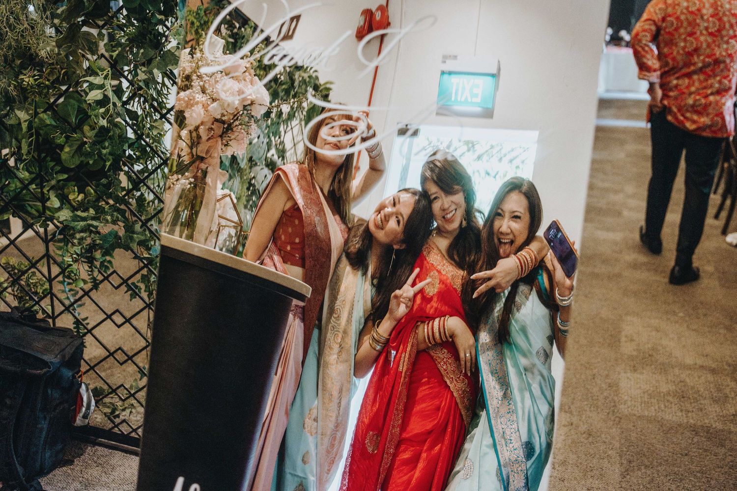 Bridesmaids in Indian wedding attire pose joyfully at a photo booth cutout display.