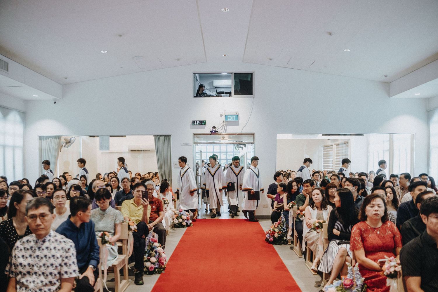 Wedding ceremony in a white church with guests seated along a red carpet aisle as the bridal party walks.