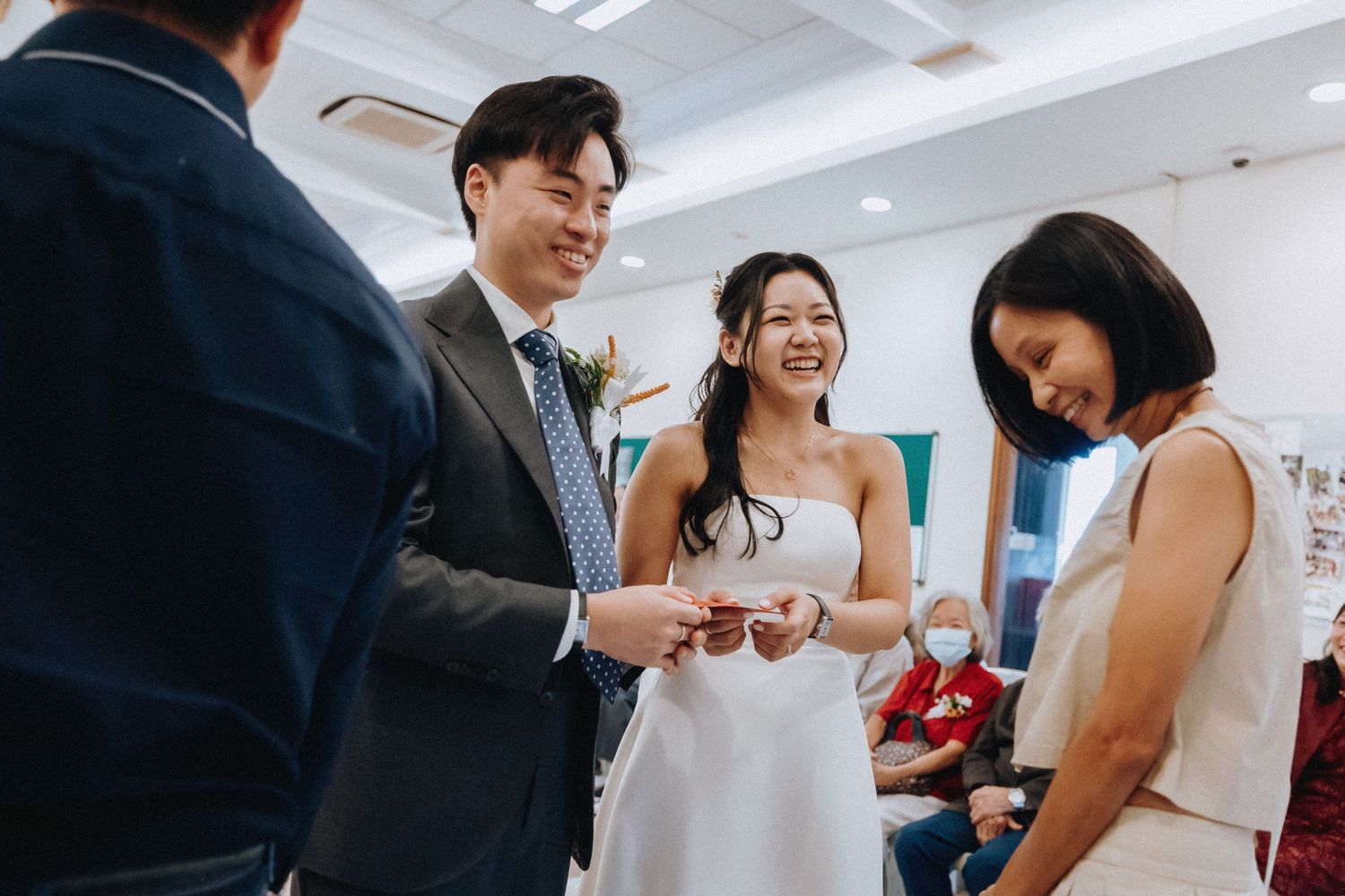 Smiling bride and groom exchange rings during intimate wedding ceremony with guests watching nearby.