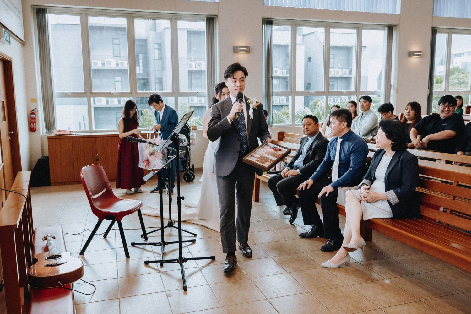 A groom delivers a heartfelt speech to seated wedding guests inside a bright church hall.