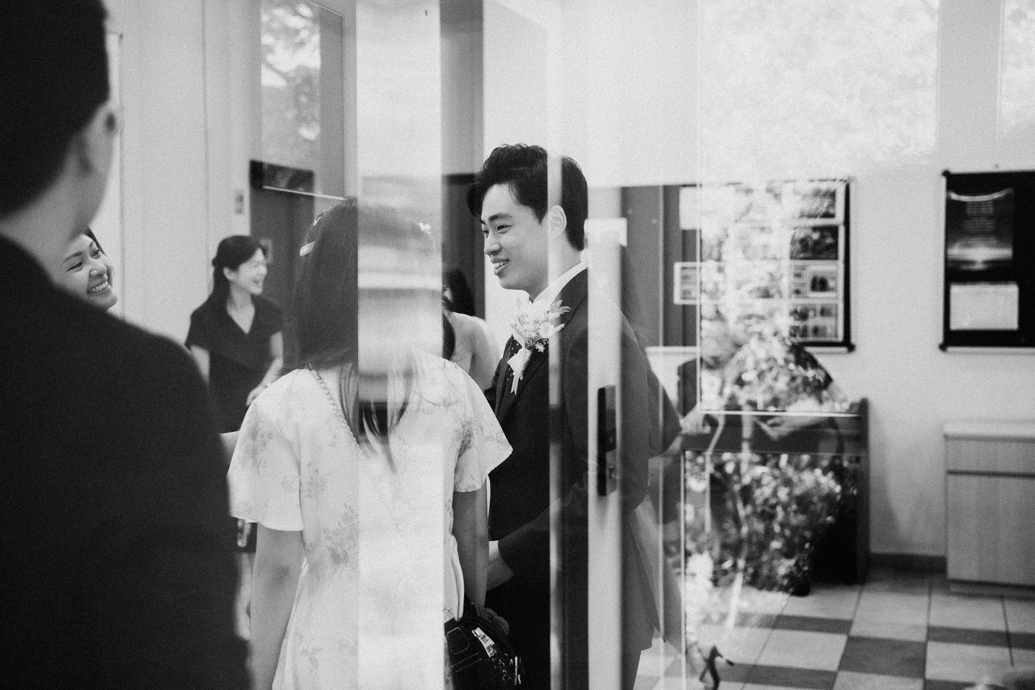 Black and white photo of smiling groom in suit greeting guests at wedding venue entrance.