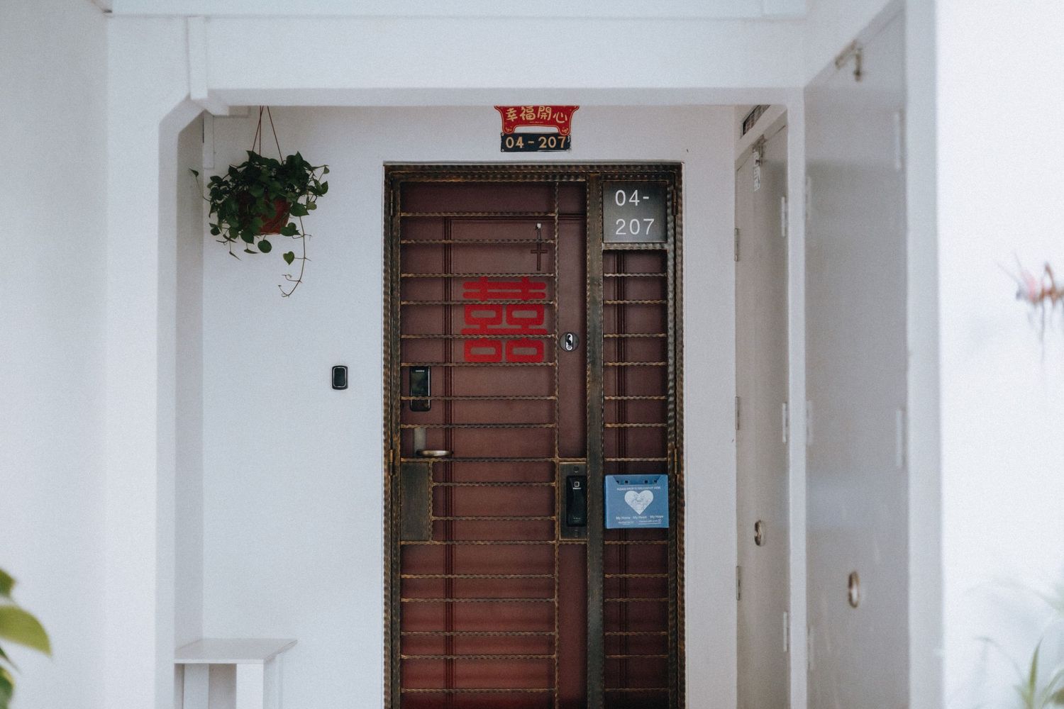 Brown wooden front door with red notices and plants in a white HDB apartment corridor in Singapore.