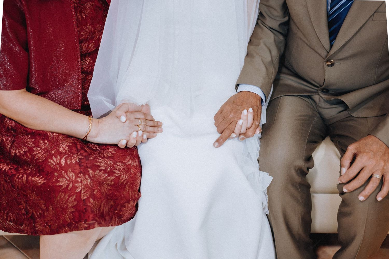 Bride in white gown seated between parents in formal attire at a wedding ceremony, holding hands.