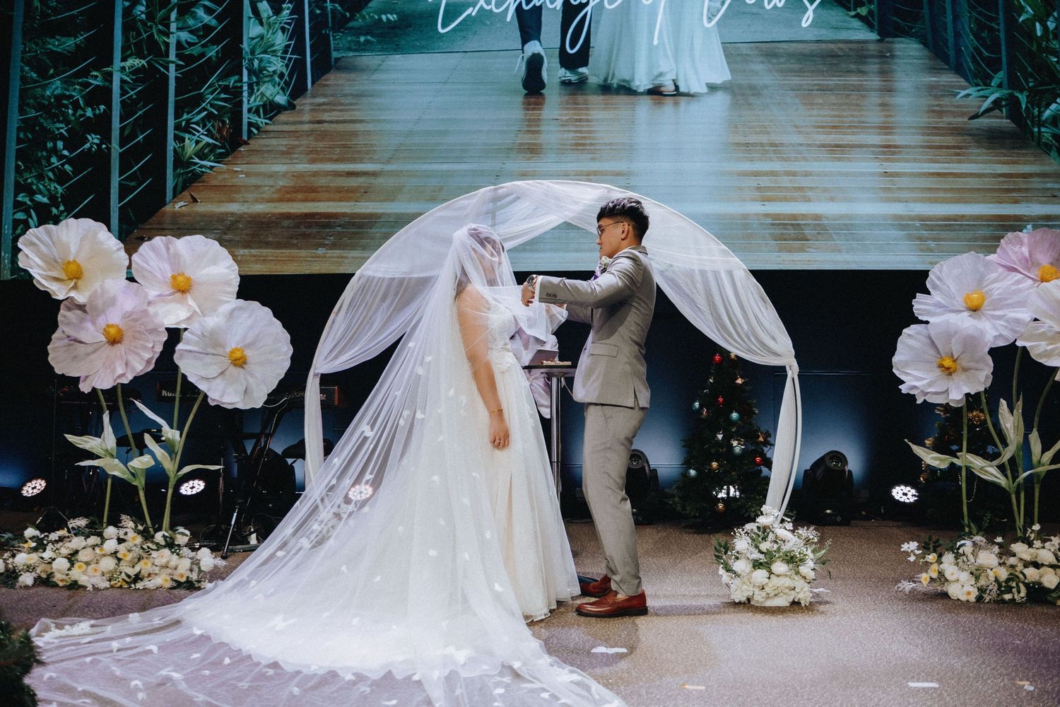 A wedding couple poses under a floral arch with white orchids and flowing veil in an elegant setting.