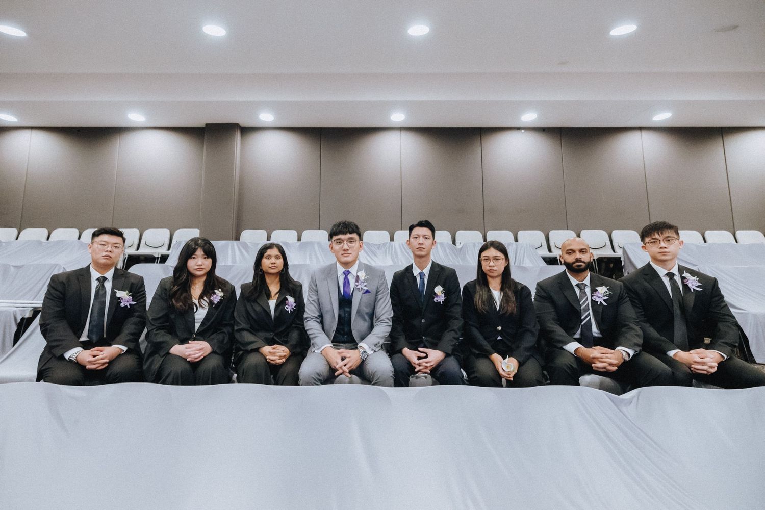 Formal group photo of wedding attendees seated in rows of auditorium chairs.