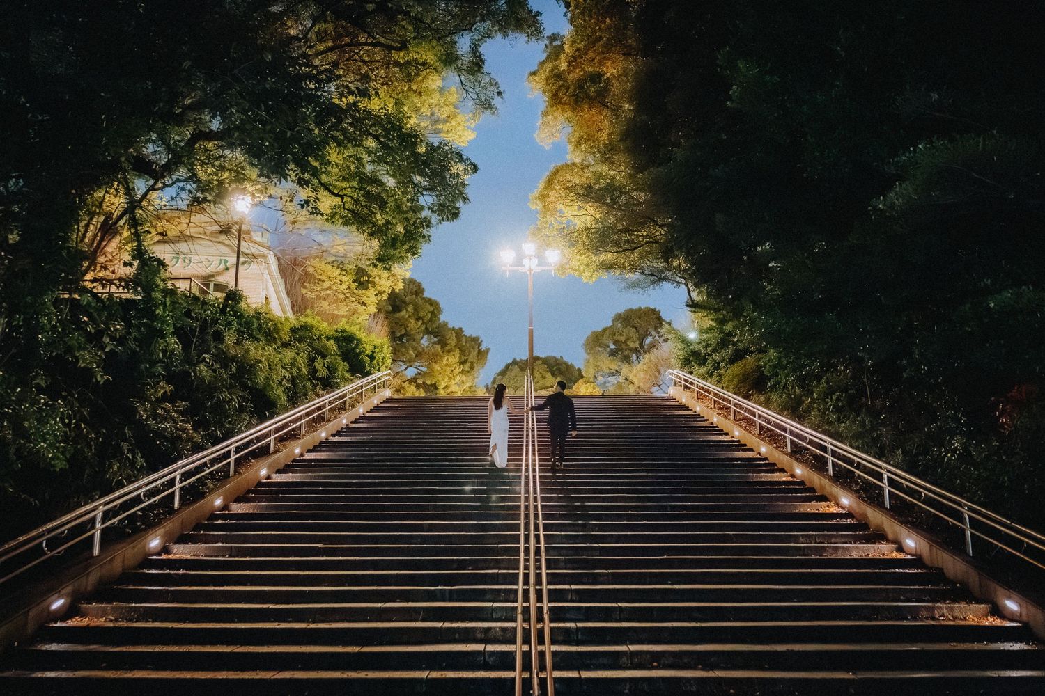 Person with umbrella ascending illuminated grand staircase flanked by lush trees under a clear blue sky.