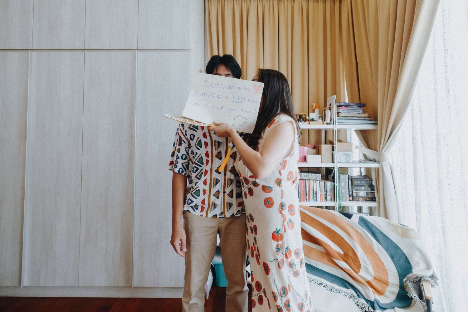 Two people in floral dresses standing in a bedroom with beige curtains and blue decor.