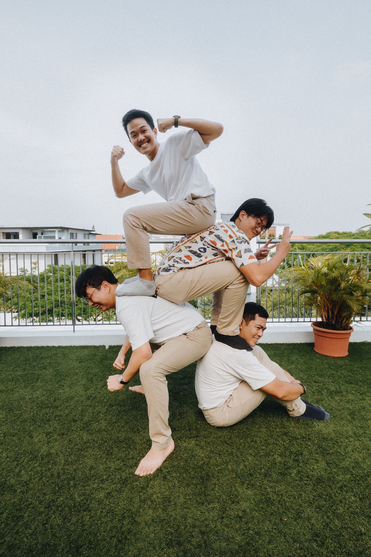 A playful group poses in a creative stacked formation on an outdoor rooftop terrace with potted plants.