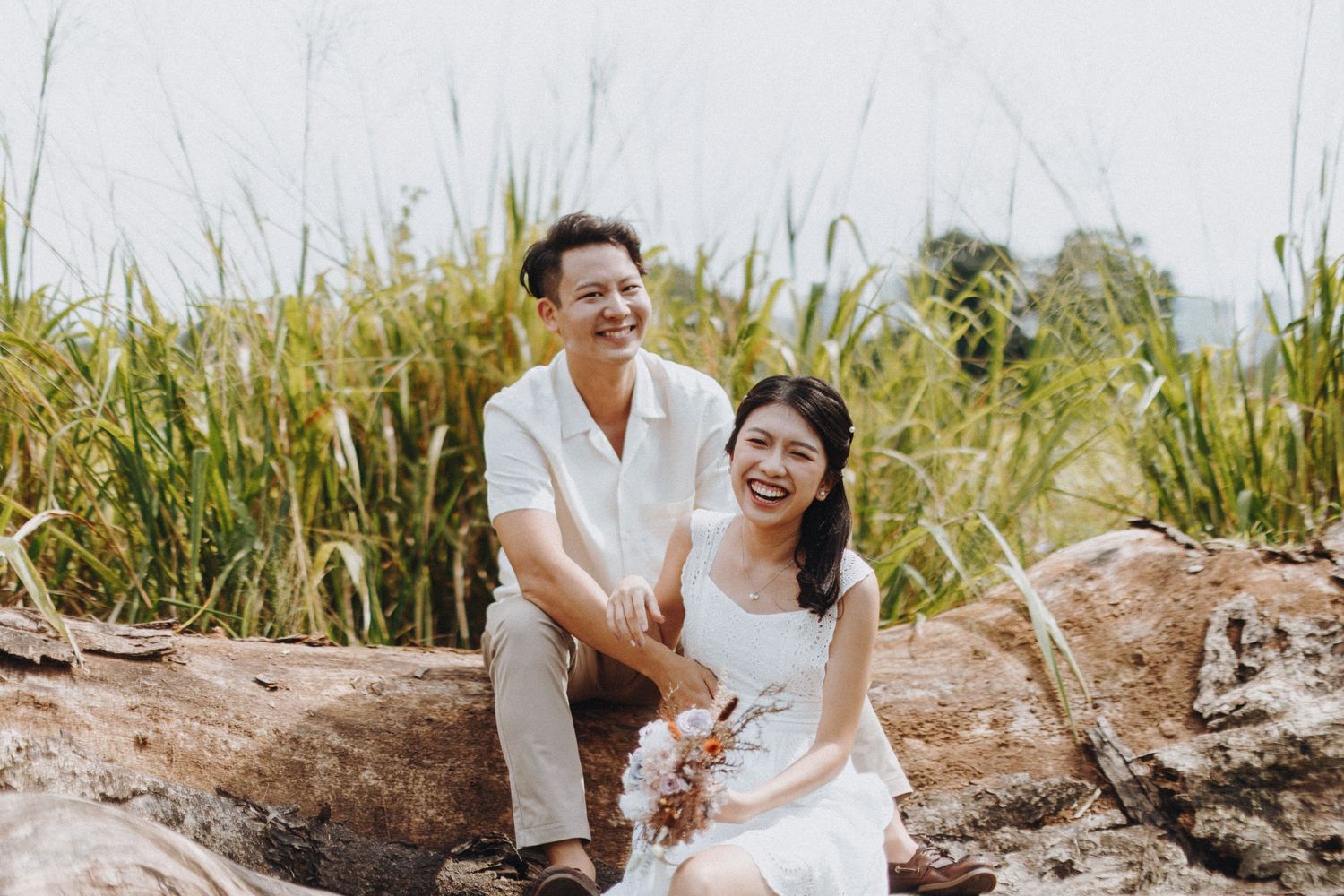 A couple sits together on a fallen log surrounded by tall grass in a natural outdoor setting.