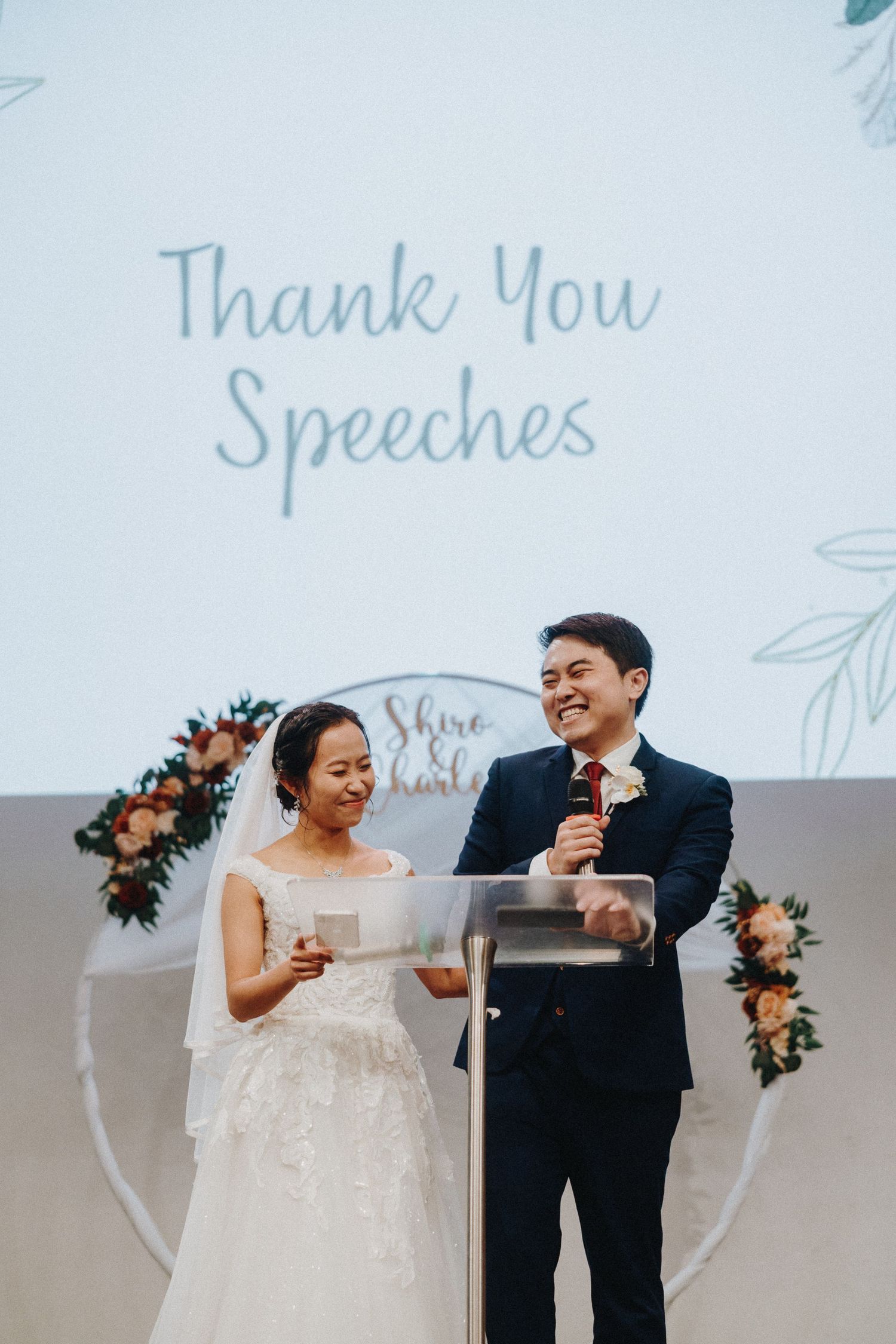 A couple delivering thank you speeches at their wedding reception with floral decorations behind them.