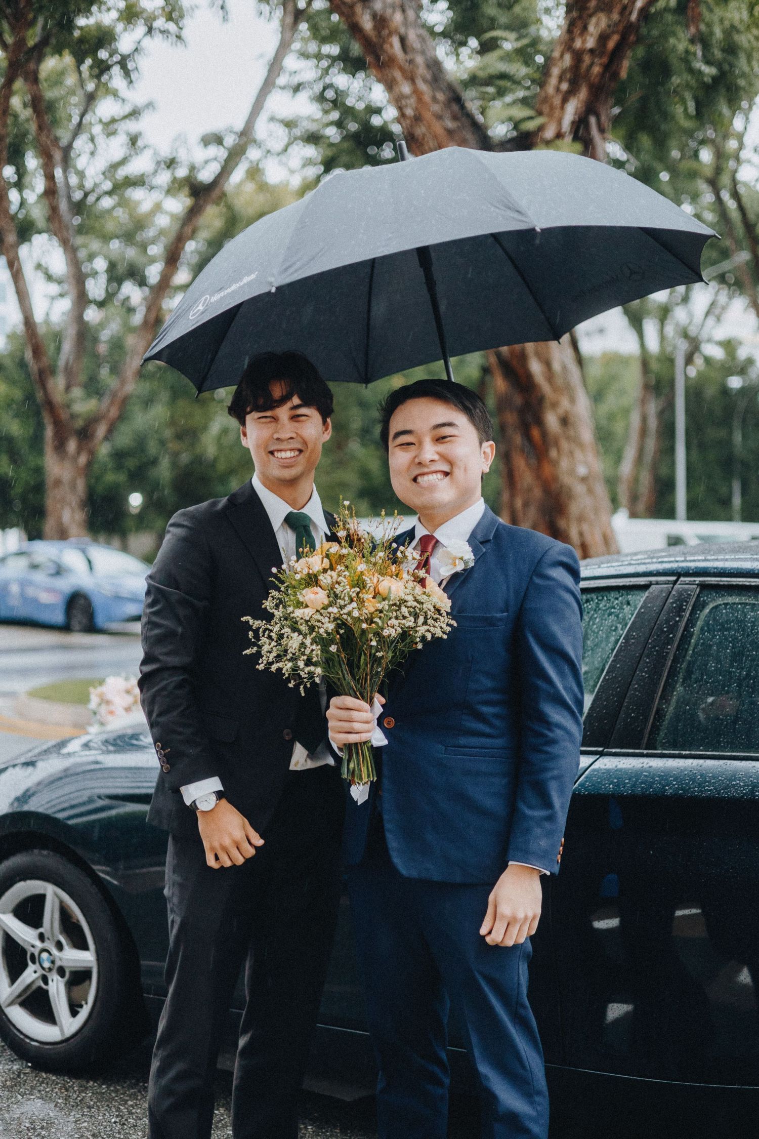 Two people in formal suits share an umbrella while holding a bouquet on a rainy day by a parked car.