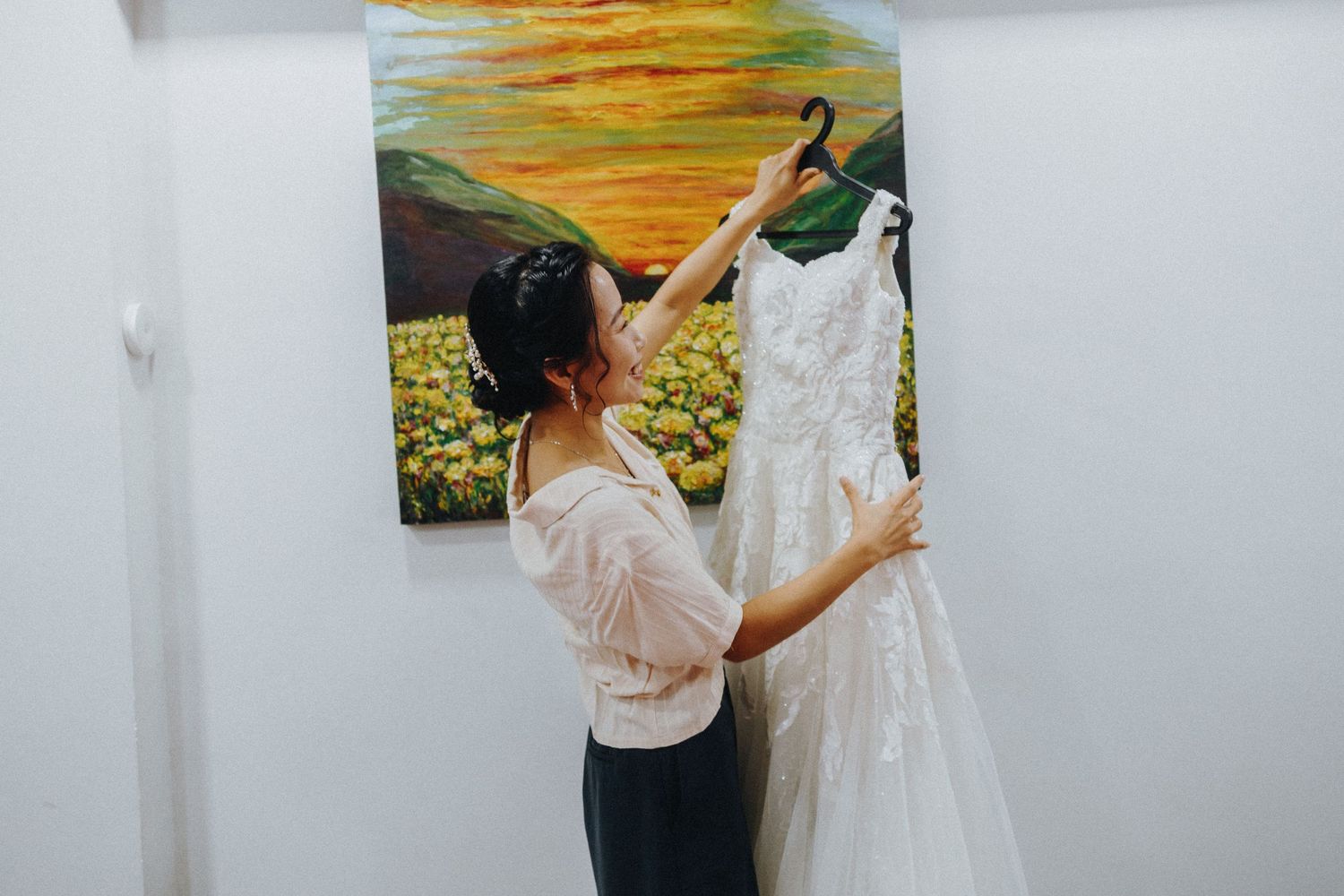 A white wedding dress being held up to examine details while hanging against a bright yellow and green painting background.