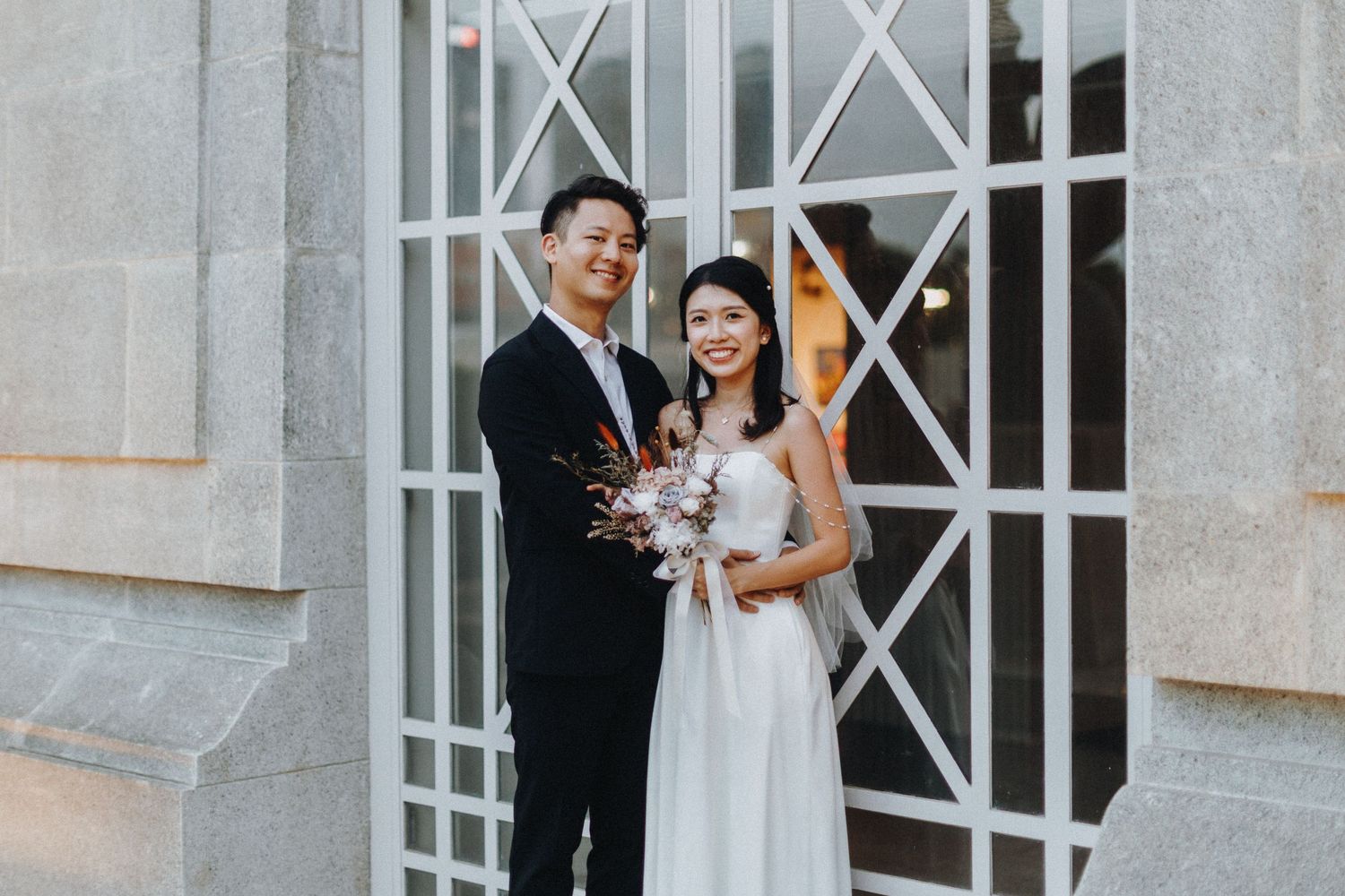 A couple in wedding attire poses in front of an ornate white gate entrance.