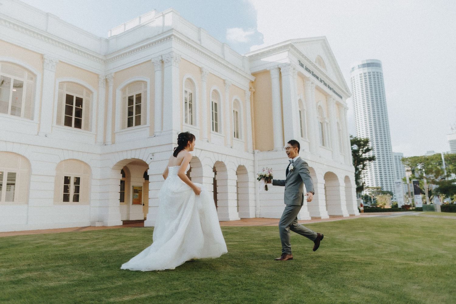 A wedding couple playfully runs on the lawn in front of a grand white colonial-style building.