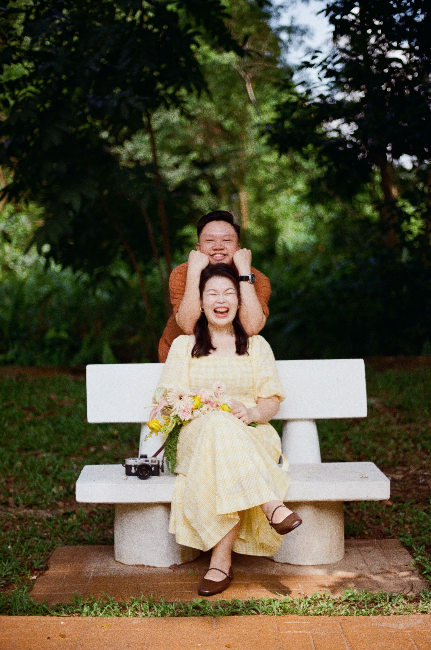 A joyful couple sharing a romantic moment on a white concrete bench surrounded by lush greenery in a park setting.